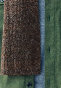 Groene wollen jas met een bruine, gestructureerde gebreide sjaal over een lichtblauwe trui. Voorzien van knoopsluiting en subtiele decoratieve stiksels.