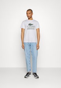 Camiseta blanca de algodón con un logotipo de cocodrilo verde bordado y el texto "LACOSTE". Combinada con unos vaqueros azules claros y zapatillas negras.