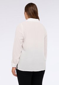 Weiße Bluse mit langen Ärmeln, feinem strukturierenden Muster, Knopfkragen und seitlichen Schlitzen am Saum, kombiniert mit schwarzen Hosen.