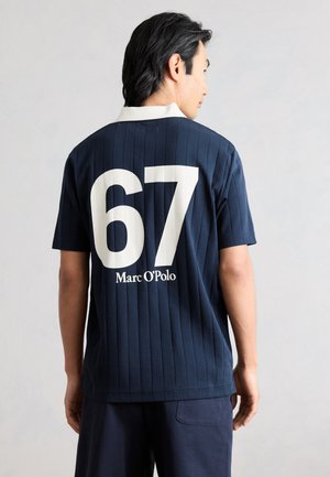 Homme de dos portant une chemise bleu marine à manches courtes avec un col blanc, un grand numéro 67 et le texte "Marc O'Polo" dans le dos.