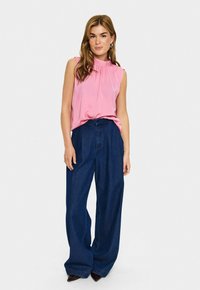 Ženska v brezrokavnem roza bluzi in širokih temno modrih hlačah, ki stoji ob enobarvni beli podlagi.