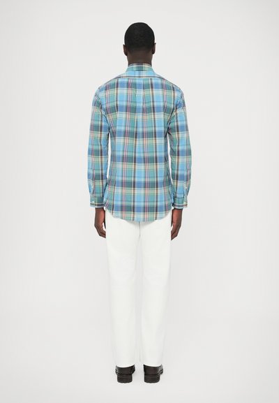 Srajca z dolgimi rokavi s tartan vzorcem v turkizni, modri in beli barvi. Ima ovratnik in manšete z gumbi. Kombinirana z belimi hlačami in črnimi čevlji.