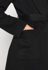 Svart kappa med bälte i midjan, vida ärmar och en stor ficka på vänster sida, tillverkad av ett texturerat tyg.