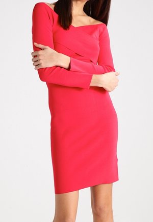 Kobieta ubrana w dopasowaną, długorękawową, jasnoróżową sukienkę do kolan z kwadratowym dekoltem, stojąca z założonymi skrzyżowanymi ramionami.