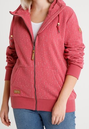 Kobieta w różowej bluzie z kapturem na zamek błyskawiczny z małymi białymi wzorami serduszek, kapturem ze sznurkiem, przednimi kieszeniami i niebieskich dżinsach.