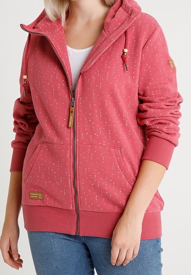 Femme portant un sweat à capuche zippé rose avec petits motifs de cœurs blancs, capuche à cordon, poches avant et jean bleu.