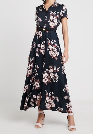 Robe longue bleu marine à motifs floraux rose et blanc, manches courtes, col, ceinture noire, boutonnée sur le devant, portée avec des talons roses clairs à bride cheville.