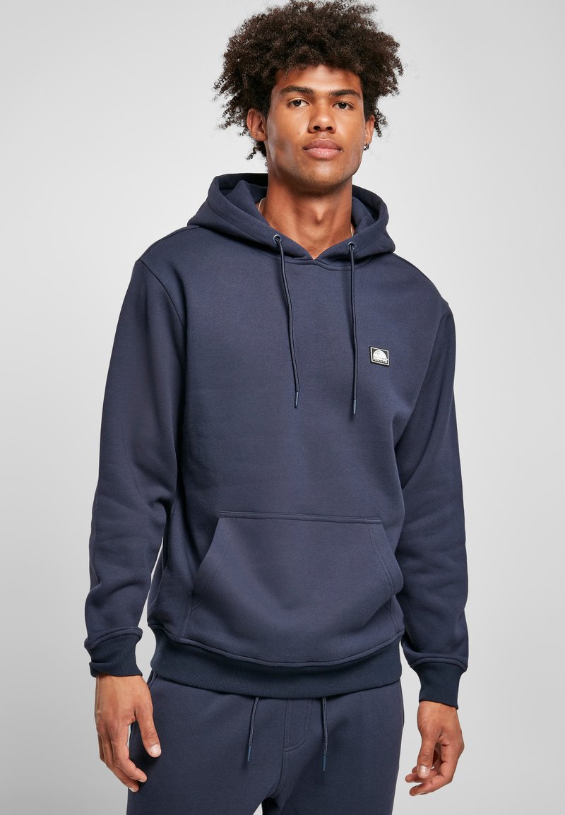 Navy blue hoodie made of soft fabric, featuring a front pocket, drawstring hood, and a small logo patch on the chest.