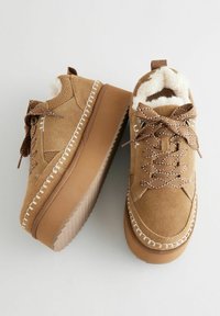 Sneakers in suede color tan con rivestimento morbido, suole spesse in beige e cuciture decorative. Lacci marroni con un motivo intrecciato.