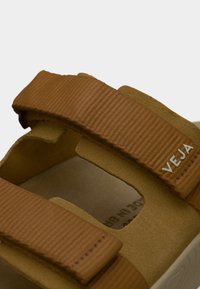 Καφέ vegan σανδάλια με δύο φαρδιές, υφασμάτινες λουριά, επενδεδυμένη βάση και ορατό λογότυπο της μάρκας. Ο σχεδιασμός διαθέτει ανοιχτή μύτη.