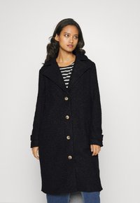 Cappotto in misto lana nera con finitura testurizzata, ampio colletto e cinque bottoni in legno. Indossato sopra una maglia a righe con maniche lunghe.