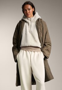 Gombolt olívazöld külső dzseki tejes színű polár kapucnis pulóver felett, fehér, bő szárú nadrággal párosítva. Magas gallérral és bordázott derékkal rendelkezik.