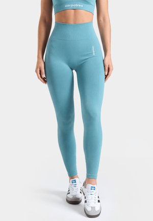 Kobieta w niebieskich legginsach z wysokim stanem i dopasowanym crop topie, stoi z rozluźnionymi rękami, nosi białe buty sportowe Adidas.