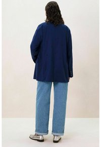 Blazer bleu marine avec un motif texturé, coupe décontractée, poches avant et poignets à trois boutons, associé à un jean baggy bleu clair et des chaussures argentées brillantes.