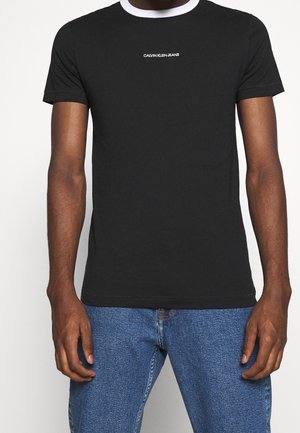 T-shirt noir Calvin Klein Jeans avec col rond blanc porté par une personne portant un jean bleu sur un fond uni.