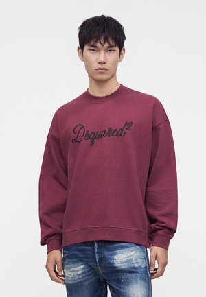 Νέος άνδρας που φοράει μπορντό φούτερ Dsquared2 και σκισμένο μπλε τζιν, στέκεται μπροστά σε απλό φόντο.