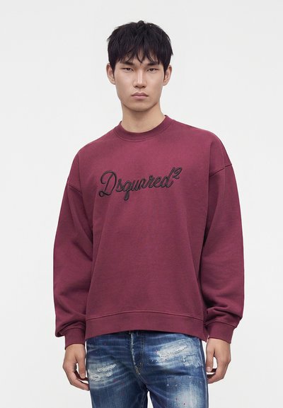 Νέος άνδρας που φοράει μπορντό φούτερ Dsquared2 και σκισμένο μπλε τζιν, στέκεται μπροστά σε απλό φόντο.