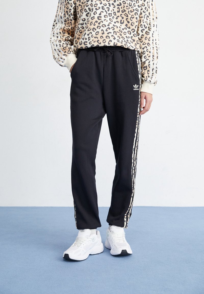 Henkilö, jolla on päällään mustat Adidas-trainingshousut, joissa on leopardikuosiset raidat, ja valkoiset lenkkarit, seisoo sinisellä lattialla valkoista seinää vasten.