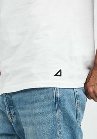 Vit t-shirt med kort ärm och svart triangellogo vid fållen, ihop med ljusblå jeans i denim med sömdetaljer.