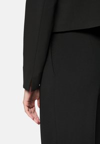 Black tailored blazer with a slightly structured design. Features clean lines, a sharp collar, and side vent detail. Smooth fabric texture.