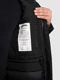 Interior de chaqueta negra con una etiqueta blanca que muestra instrucciones de cuidado, bolsillo con cremallera y forro acolchado. Textura suave en general.