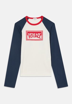 Chemise à manches longues avec manches bleu marine, corps blanc, col rouge et logo Versace rouge centré sur la poitrine dans une bordure géométrique.