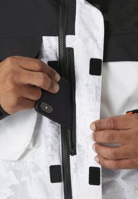 Chaqueta en blanco y negro con cremallera, que presenta un patrón texturizado. Una mano coloca un teléfono inteligente negro en un bolsillo interior.