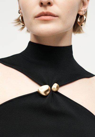 Γυναίκα που φοράει μαύρο τοπ με ψηλό γιακά και άνοιγμα κλειστό με δύο ακανόνιστα χρυσά κουμπιά, και χρυσά κρίκους σκουλαρίκια, ουδέτερο φόντο.