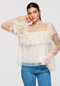 Jeune femme aux cheveux foncés portant un chemisier en dentelle blanche transparente et un jean bleu, reposant sa tête sur sa main devant un fond clair.