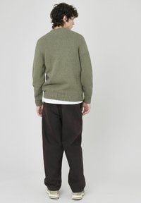 Pull tricoté vert olive avec un tissu texturé, col rond et ourlet côtelé, associé à un pantalon noir ample et des chaussures de couleur claire.