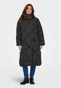 Ilgas juodas pagalvę primenantis pūkinis paltas su aukštu apykakle, su sagų užsegimu ir deimantuota siuvinėjimu; dėvimas ant mėlynų džinsų ir juodų kulkšninių batų.