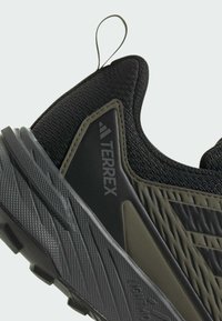 Schwarzer Sportschuh mit strukturiertem Obermaterial, grünen Akzenten und einer Gummisohle mit strukturiertem Profil. Reflektiertes Logo an der Seite.