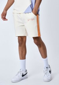 Pantalones cortos de crema para hombre con franjas laterales naranjas, fabricados en un tejido suave. Cuentan con una cintura ajustable con cordón y bolsillos laterales. Llevados con calcetines y zapatos blancos.