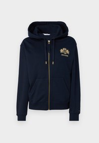 Námornícky modrá zip-svetrová mikina vyrobená z mäkkého materiálu, s vyšívaným zlatým logom, dvoma prednými vreckami a kapucňou na šnurovanie.