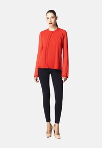 Maglietta rossa a maniche lunghe con design plissettato sul davanti, abbinata a leggings neri e scarpe con tacco beige. Tessuto liscio con silhouette aderente.