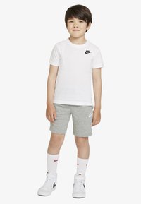 Maglietta bianca a maniche corte Nike con logo nero, pantaloncini di cotone grigi, sneakers alte bianche e calzini bianchi con dettagli rossi.