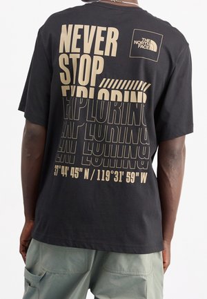 Henkilö pukeutuneena mustaan t-paitaan, jossa on beige teksti "Never Stop Exploring" ja The North Face -logo, yhdistettynä vihreisiin housuihin.