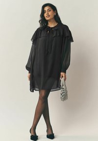 Fekete, áttetsző ruha fodros dekoltázzsal és hosszú, puffos ujjal. Ezüst szőtt táskával és fekete, hegyes orrú balerina cipővel párosítva.