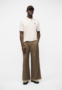 Άνδρας με μπεζ πόλο πουκάμισο, καφέ παντελόνι με φαρδιά μπατζάκια, μαύρα παπούτσια και καφέ καπέλο, στέκεται με το ένα χέρι στην τσέπη σε λευκό φόντο.