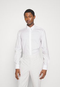 Weißes Langarmhemd mit Button-Down-Kragen, aus glattem Stoff, kombiniert mit hellgrauen, strukturierten Hosen mit Knopfverschluss.