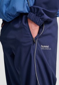 Marinblå träningbyxor med en dragkedjeficka, texturerad tyg, elastisk midja och ljusgrå pipingdetaljer. Hummel-logotypen är synlig.