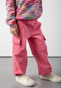 Dziecko w jasnoróżowych spodniach cargo, wielokolorowym swetrze robionym na drutach i białych sneakersach stoi na szarej podłodze przy białej ścianie.