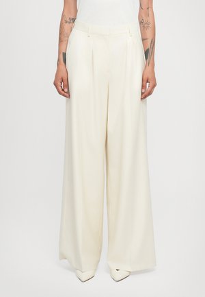 Wide-leg trousers in lightweight cream fabric, featuring front pleats, belt loops, and a tailored fit, paired with pointed white shoes.