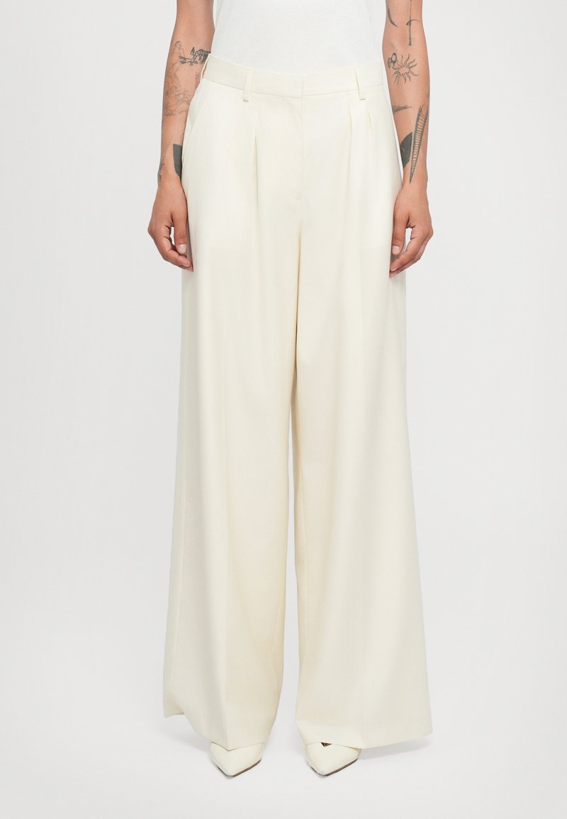Wide-leg trousers in lightweight cream fabric, featuring front pleats, belt loops, and a tailored fit, paired with pointed white shoes.