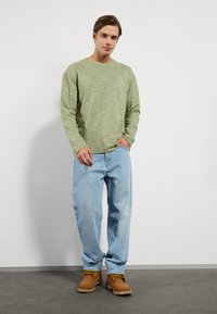 Chemise longue à manches longues texturée verte, jean décontracté bleu clair, et bottines brunes à lacets jaunes. Debout sur un sol gris.