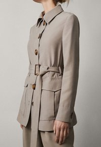 Jasnokremowa kurtka zapinana na pasek z wyraźnym kołnierzem, dużymi guzikami i przednimi kieszeniami, wykonana z gładkiego, tkanej tkaniny o dopasowanym kroju.