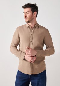 Camicia button-up a maniche lunghe color marrone chiaro con una texture morbida e colletto a punta, abbinata a jeans blu scuro.