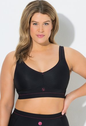 Femme portant un soutien-gorge de sport noir avec des coutures roses et un legging taille haute assorti, debout avec une main sur la hanche devant un fond blanc.