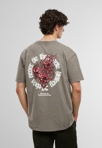Tricou oversized gri cu mâneci scurte, având un grafic roșu cu un craniu și flăcări pe spate, și text alb în jurul design-ului.