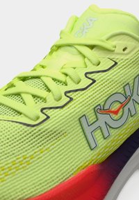 Αθλητικό παπούτσι σε λαχανί πράσινο με πλεκτό επάνω μέρος, επίπεδα κορδόνια και μαλακή σόλα που μεταβαίνει από κόκκινο σε μοβ. Διαθέτει λογότυπο στην πλευρά.